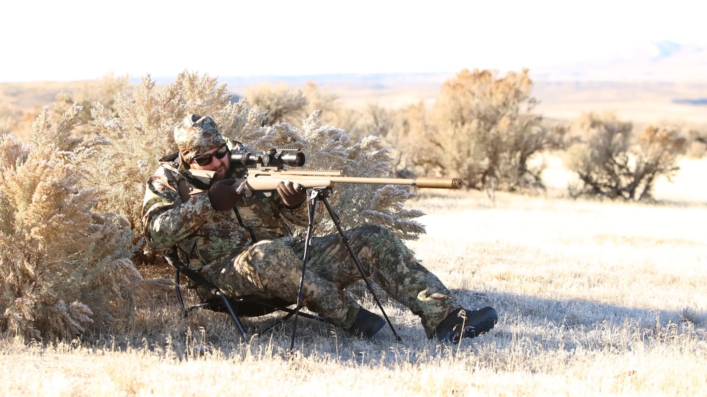 A hunter in camouflage sitting low against a frost-covered sagebrush, shouldered behind a suppressed bolt-action rifle resting on a bipod, glassing open desert country beyond.